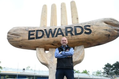 Ieuan Edwards poses with his arms crossed in front of a giant artificial fork with a giant artificial sausage with the word 'Edwards' engraved into it