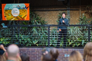 Ellen MacArthur, founder of the EMF, stands with a microphone in her hand facing an audience of people, stood next to a billboard which reads 'Big Food Redesign Challenge'