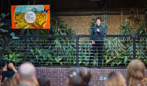 Ellen MacArthur, founder of the EMF, stands with a microphone in her hand facing an audience of people, stood next to a billboard which reads 'Big Food Redesign Challenge'