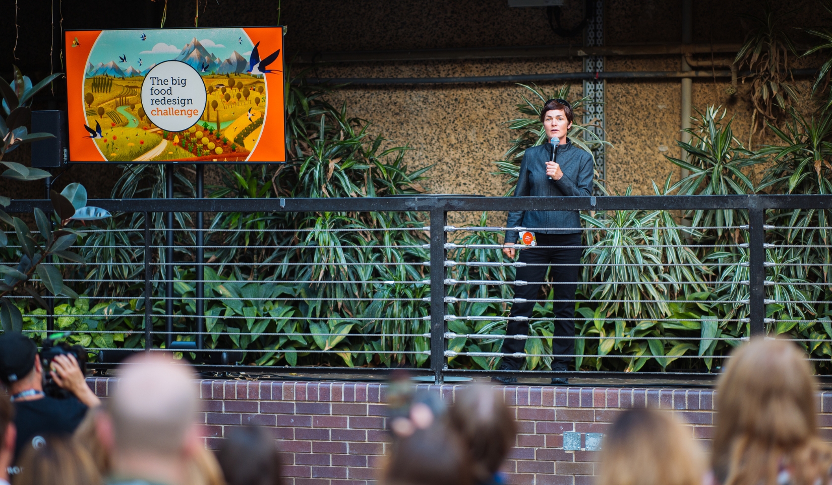 Ellen MacArthur, founder of the EMF, stands with a microphone in her hand facing an audience of people, stood next to a billboard which reads 'Big Food Redesign Challenge'